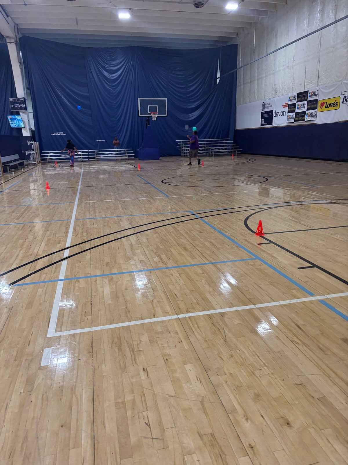 Empty gymnasium with wooden floor, blue curtain backdrop, basketball hoop, and red training cones set up for drills