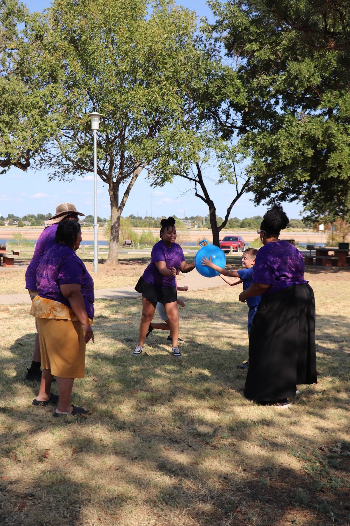Four women in purple shirts playing frisbee outdoors under large shade trees in a rural park setting
